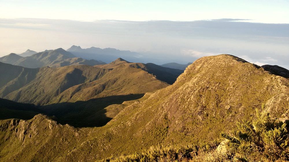 O Pico do Calçado iluminado pelo sol, visto do alto do Pico da Bandeira, com a serra ao fundo