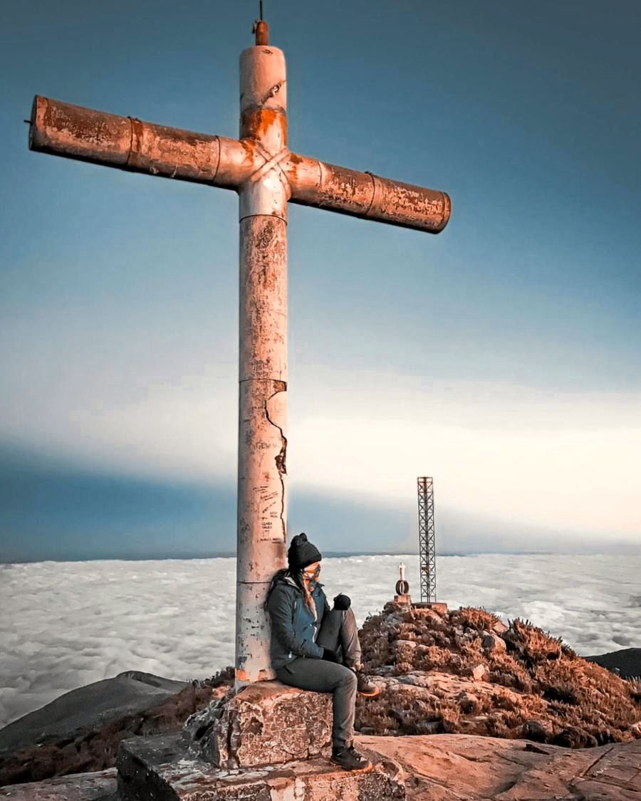 Visitante sentada junto ao cruzeiro do cume, com a torre metálica e um mar de nuvens ao fundo
