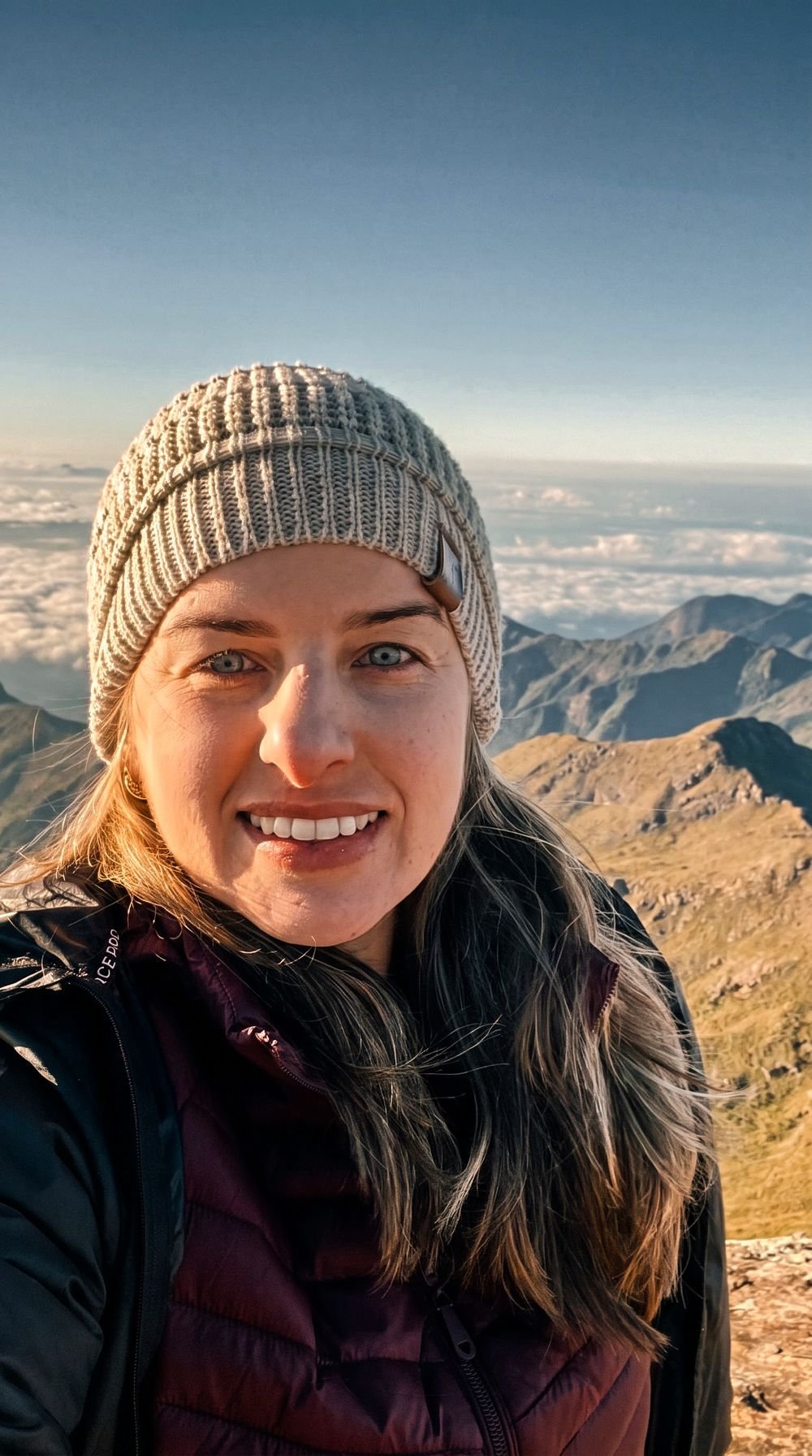 Montanhista de gorro e jaqueta no alto da serra, sorrindo, com o mar de nuvens e as cristas ao fundo