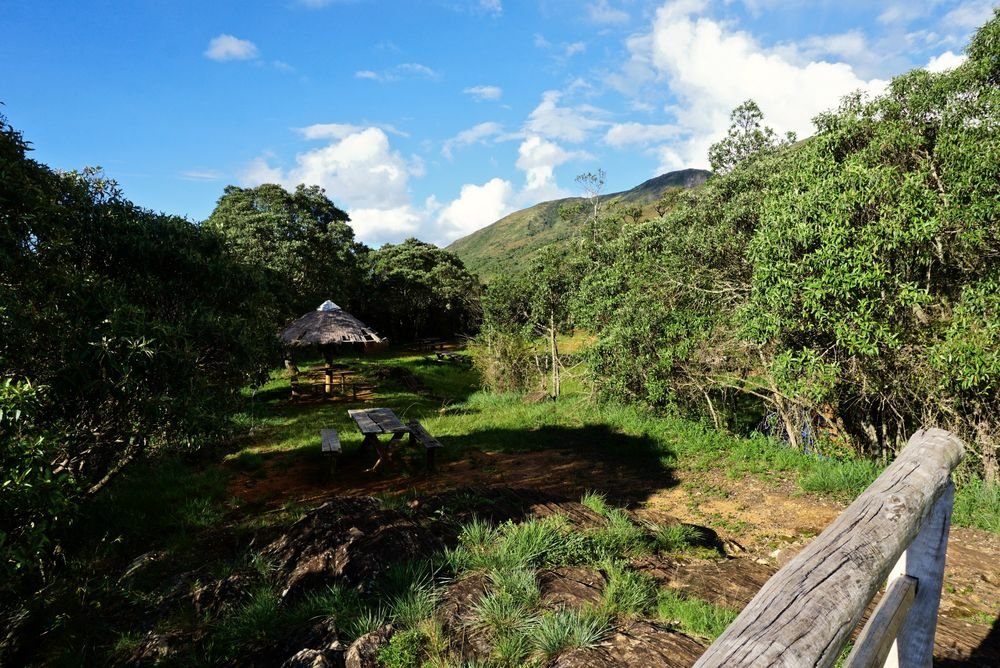 Área de acampamento da Tronqueira, com quiosque de palha, mesas e cerca de madeira, sob o céu azul