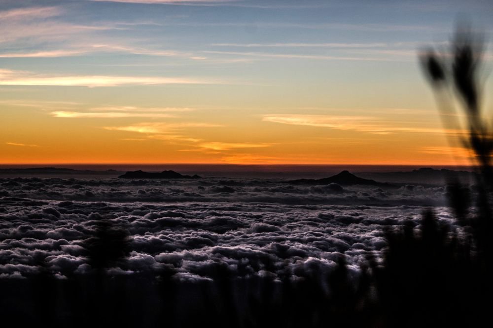 O céu alaranjado do início da alvorada sobre o mar de nuvens, visto do alto do Pico da Bandeira