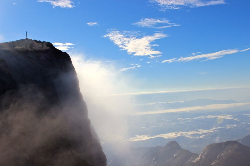 O paredão do Pico da Bandeira de perfil, com o cruzeiro no alto e a névoa subindo pela encosta
