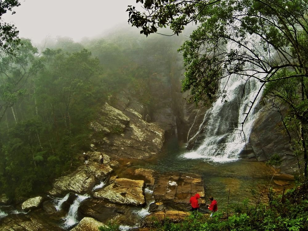 Cachoeira descendo entre as pedras e a mata, com névoa entre as árvores e duas pessoas de vermelho ao pé da queda