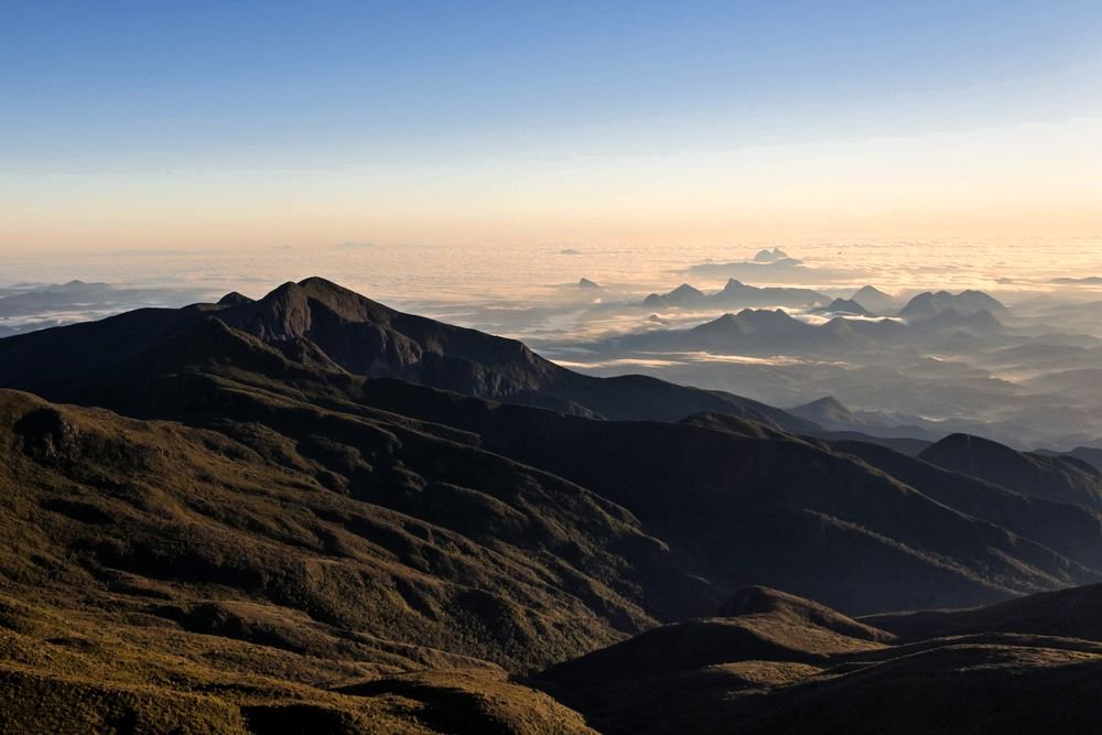 Cristas escuras da serra ao amanhecer, com o mar de nuvens e picos distantes no horizonte