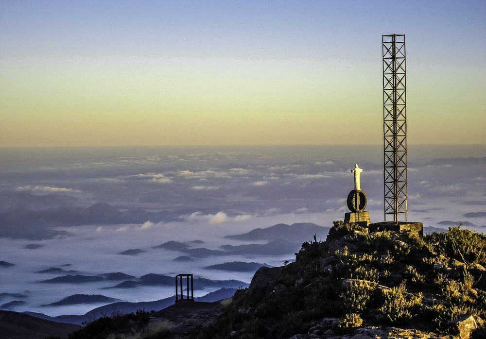 A estátua do Cristo e a torre metálica no cume do Pico da Bandeira, acima do mar de nuvens