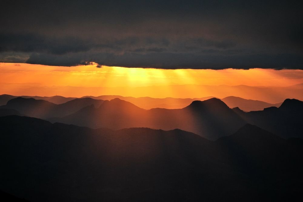 Raios de sol alaranjados atravessando as nuvens escuras sobre as montanhas