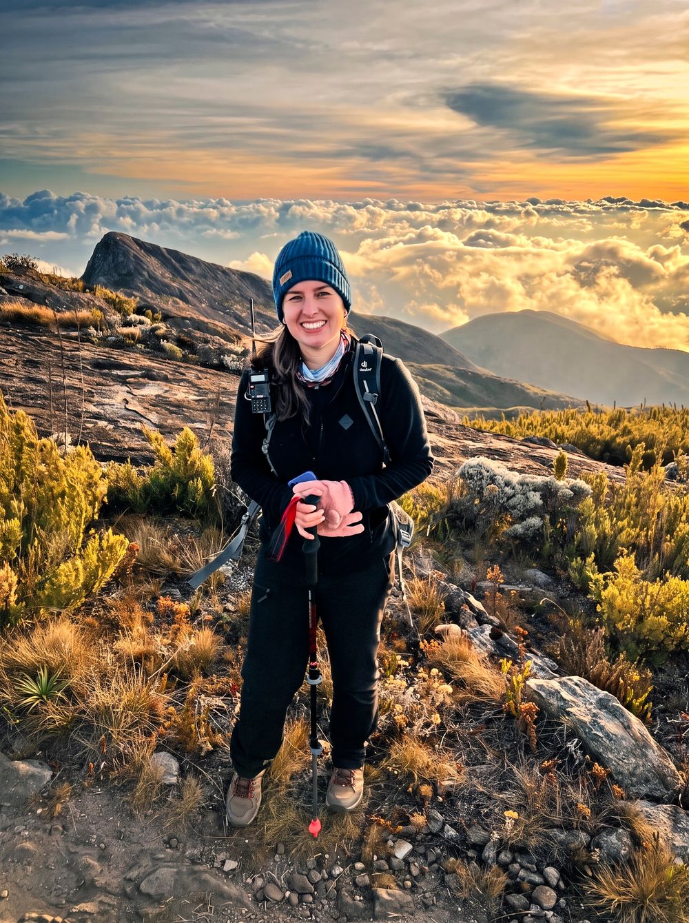 Kely Abreu no alto da serra ao nascer do sol, com gorro, rádio e bastão de caminhada, diante do mar de nuvens