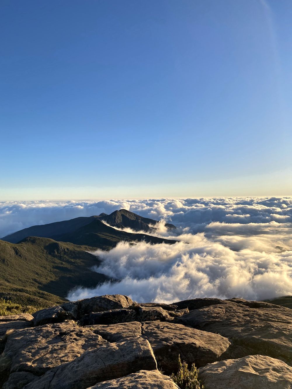 Mar de nuvens sob o céu azul, visto do alto da serra, com rochas em primeiro plano