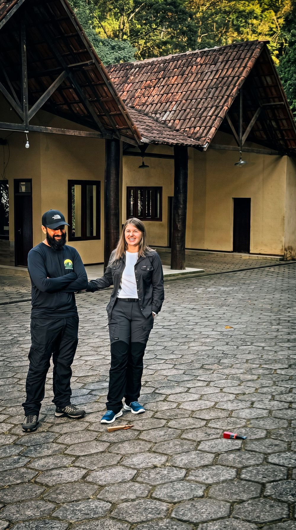 Sairo e Kely sorriem no pátio de pedra da portaria do parque em Alto Caparaó, diante da construção de telhado de telhas e madeira